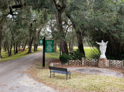 A gravel path leads to a garden with a statue and a bench, surrounded by trees and a signpost.