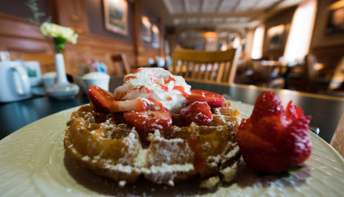 A close-up of a waffle topped with strawberries and whipped cream, served on a plate in a cozy restaurant setting.