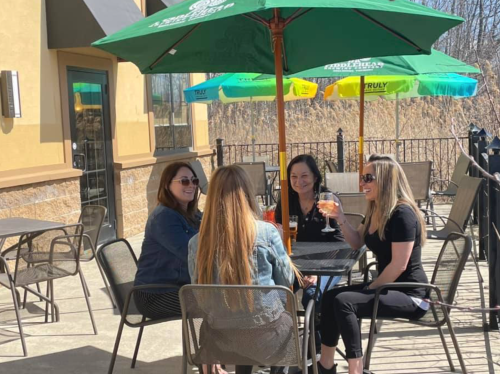 Four women sit at a table under umbrellas on a patio, enjoying drinks and chatting in a sunny outdoor setting.