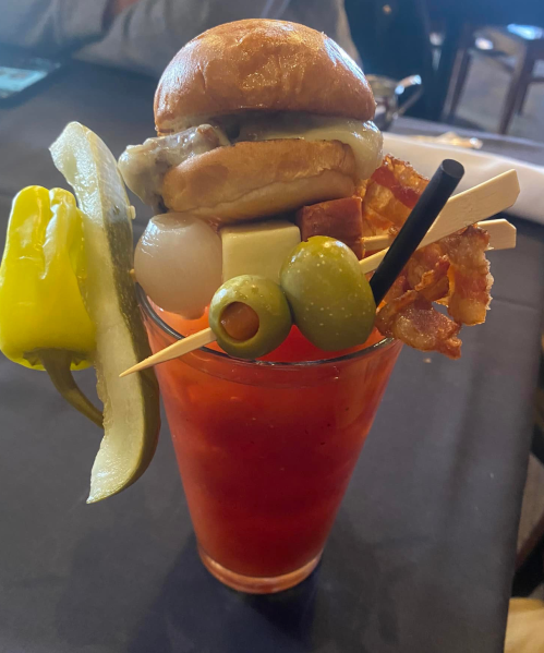 A tall cocktail glass filled with a Bloody Mary, topped with a mini burger, pickles, olives, and bacon.