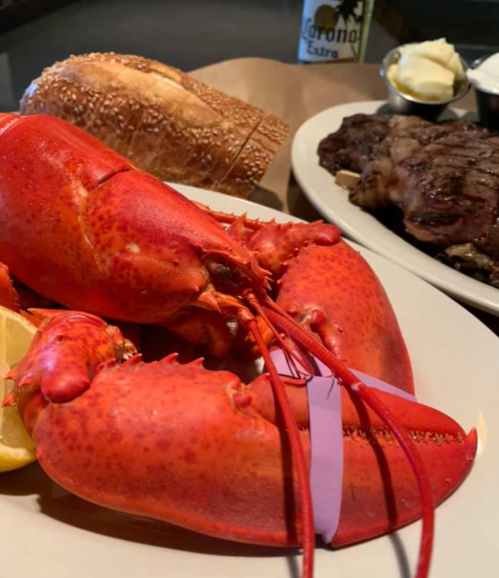A vibrant red lobster on a plate with lemon, accompanied by a roll and a steak in the background.