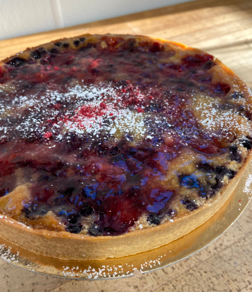 A round berry tart with a glossy fruit glaze and powdered sugar on top, resting on a wooden surface.