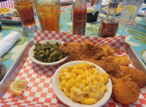 A tray with fried fish, macaroni and cheese, collard greens, and cornbread, with drinks and condiments in the background.