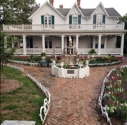 A charming white Victorian house with green shutters, surrounded by a brick pathway and colorful flower beds.