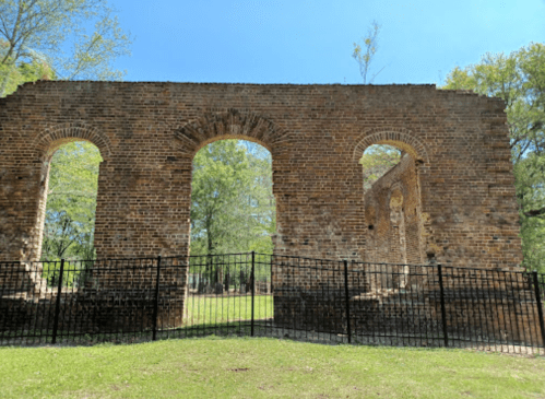Ruins of a brick structure with arched openings, surrounded by a black fence and green trees under a clear blue sky.