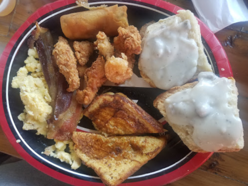 A plate of breakfast featuring scrambled eggs, fried chicken, bacon, French toast, and biscuits with gravy.
