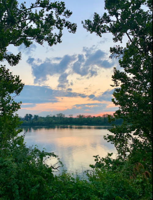 A serene lake at sunset, framed by trees, with colorful clouds reflecting on the water's surface.