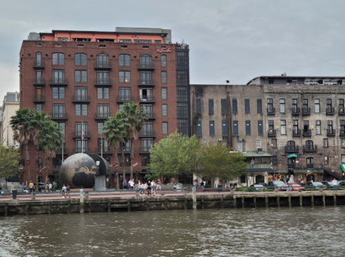 A riverside view of a brick building with balconies, palm trees, and a large globe sculpture in front.