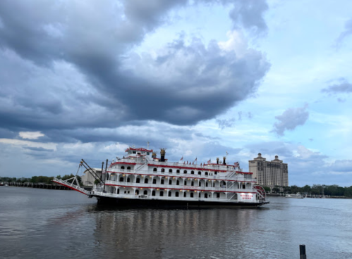 A large riverboat with red accents sails on a calm waterway under a cloudy sky, with a building in the background.