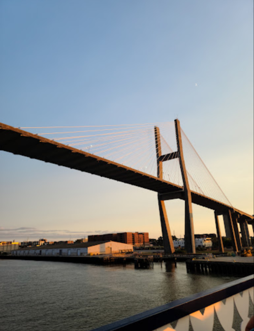 A suspension bridge spans over a river at sunset, with buildings visible along the shore.