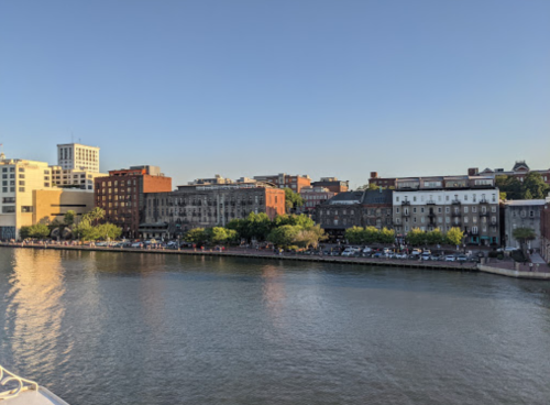 View of a calm river with buildings and trees along the waterfront under a clear blue sky.