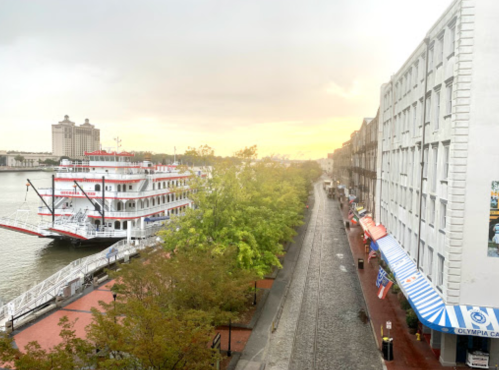 View of a riverfront with a steamboat docked, lined with trees and buildings under a cloudy sky at sunset.