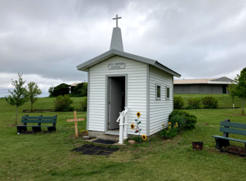A small white chapel with a cross on top, surrounded by benches and sunflowers in a grassy area.