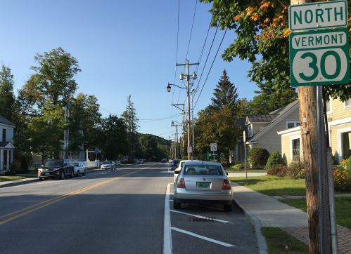 A quiet street in Vermont with parked cars, trees, and a sign for North Vermont Route 30.