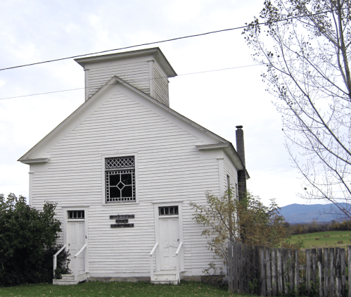 A white, historic building with a bell tower, featuring two doors and surrounded by greenery and mountains in the background.
