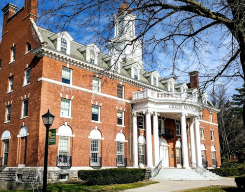 Historic brick building with white columns, surrounded by trees and a clear blue sky.