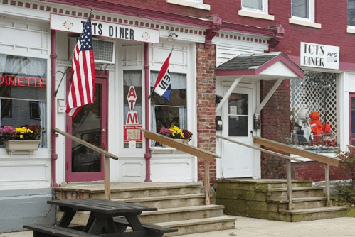 A diner with an American flag, ATM sign, and colorful flower pots, featuring a red brick exterior and wooden steps.