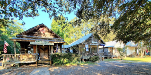 Three rustic wooden buildings with porches, surrounded by trees and greenery, in a sunny outdoor setting.