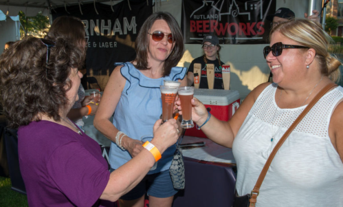 Three women toast with glasses of beer at an outdoor event, smiling and enjoying the atmosphere.