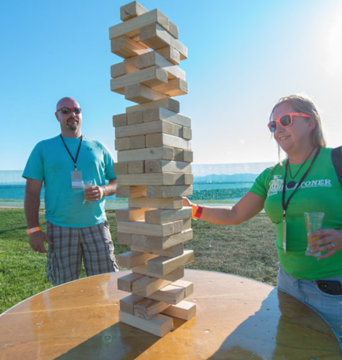 Two people play a giant Jenga game outdoors, with a tall tower of wooden blocks and a scenic background.