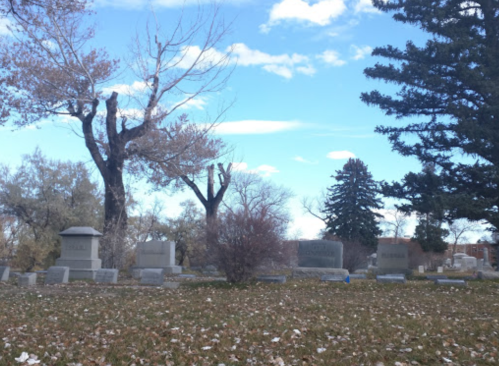 A serene cemetery scene with gravestones, bare trees, and a blue sky scattered with clouds.