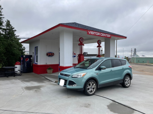 A teal SUV parked in front of a visitor center with a red roof and gas pumps, under a cloudy sky.