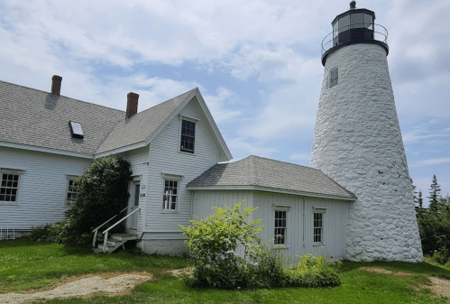 A white lighthouse beside a small, white building, set against a cloudy sky and green grass.