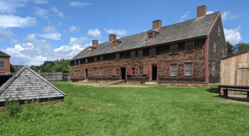Historic wooden building with multiple windows, surrounded by green grass and a blue sky with clouds.