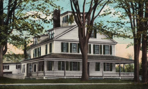 A vintage-style house with multiple stories, large windows, and a porch, surrounded by trees and a clear sky.