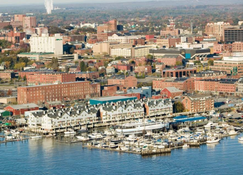 Aerial view of a waterfront city with marinas, buildings, and distant hills under a clear sky.