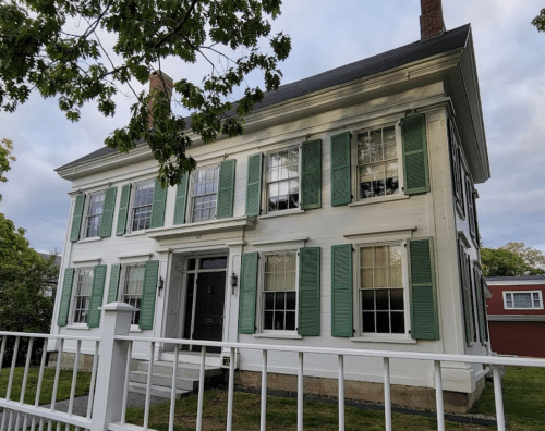 A two-story white house with green shutters and a fenced yard, set against a cloudy sky.