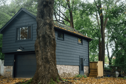 A modern dark gray house surrounded by trees, with a wooden deck and autumn leaves scattered on the ground.