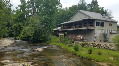 A rustic building by a calm river, surrounded by lush greenery and outdoor seating with umbrellas.