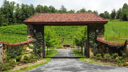 A scenic entrance gate with a stone archway, leading to a gravel driveway surrounded by lush greenery and trees.