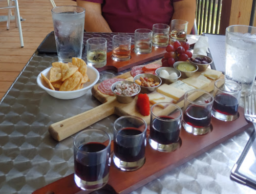 A wooden platter with wine samples, cheese, meats, nuts, and grapes, alongside a bowl of chips and glasses of water.