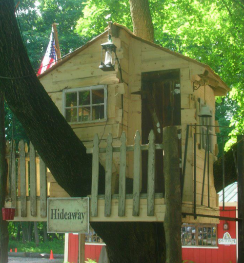 A wooden treehouse with a lantern, flag, and a sign reading "Hideaway," nestled in a lush green tree.