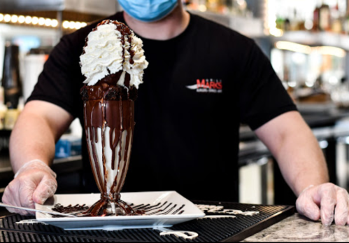 A person in a black shirt holds a tall dessert glass filled with ice cream, chocolate sauce, and whipped cream.