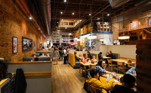 A bustling restaurant interior with brick walls, wooden floors, and diners enjoying meals at various tables.