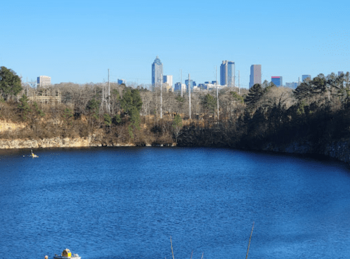 A serene lake with a city skyline in the background, featuring clear blue water and trees along the shore.