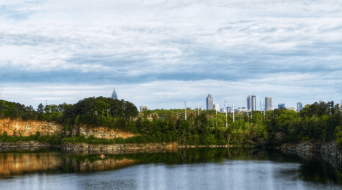 A serene lake reflects a skyline with trees and cliffs in the foreground under a cloudy sky.