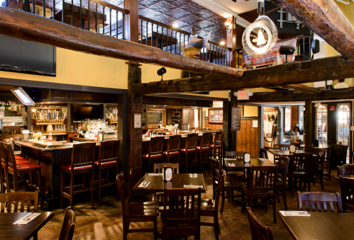 Cozy restaurant interior with wooden beams, bar area, and tables set for dining. Warm lighting creates an inviting atmosphere.