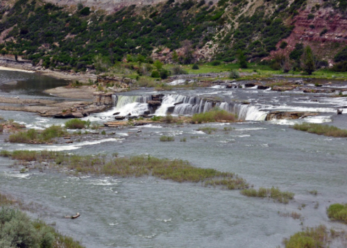 A scenic view of a waterfall cascading over rocks into a river, surrounded by greenery and rocky terrain.