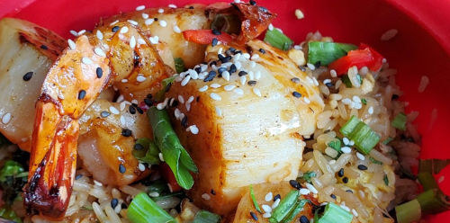 A close-up of shrimp and scallops on a bed of fried rice, garnished with green onions and sesame seeds in a red bowl.