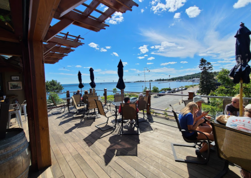 A sunny outdoor patio with chairs and umbrellas overlooking a lake and blue sky, with people enjoying the view.