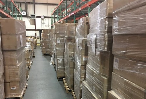 A warehouse aisle lined with tall stacks of wrapped boxes on pallets, with shelves in the background.