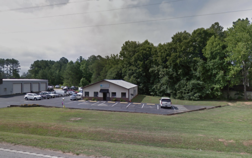 A single-story building with a parking lot, surrounded by trees, located along a road.