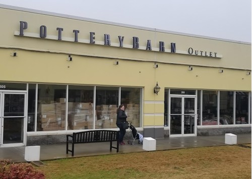 A woman stands outside a Pottery Barn Outlet store, with boxes visible through the large windows on a rainy day.