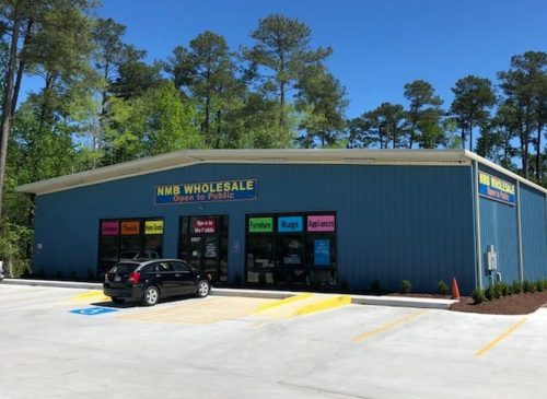 A blue warehouse-style store with "NMB Wholesale" signage, surrounded by trees and a parking lot. Open to the public.