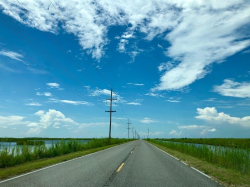 A straight road lined with power poles, flanked by water and tall grass under a blue sky with scattered clouds.
