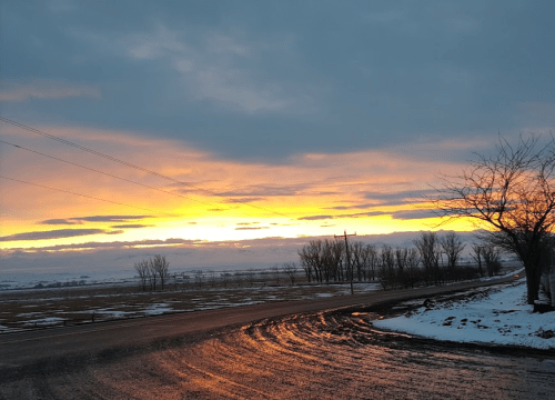 A winter sunset casts orange and pink hues over a snowy landscape, with bare trees lining a winding road.